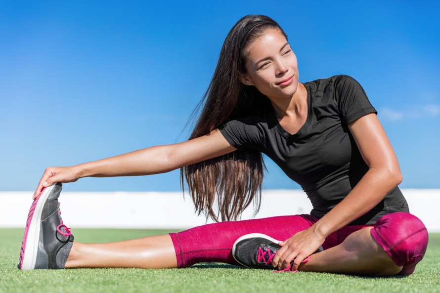 Fitness woman stretching one leg toe-touch stretch exercising hamstring and glute muscles stretches. Sporty Asian athlete exercising sitting forward bend legs exercise on grass in sunny outdoor gym.