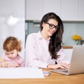 Working mother concept. Young woman working on laptop with her child from home
