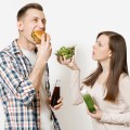 Couple man and woman standing with green detox smoothies, salad in glass bowl, burger, cola in glass bottle isolated on white background. Proper nutrition, healthy lifestyle, fast food, choice concept